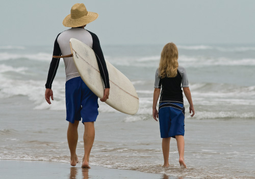 adult with hat and surfboard and child on beach walking towards the water