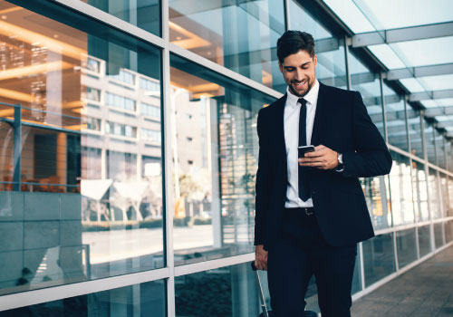 Young adult male professional walking along a modern glass building looking at his smartphone and smiling